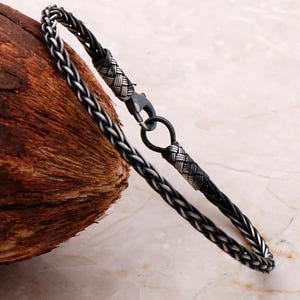 May include: A bracelet featuring a braided black cord intertwined with oxidized silver wire. The design includes two woven silver end caps and a dark metal lobster claw clasp that connects to a circular silver ring, creating a secure closure.