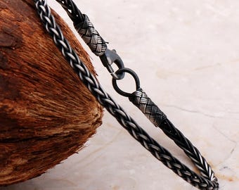 May include: A bracelet featuring a braided black cord intertwined with oxidized silver wire. The design includes two woven silver end caps and a dark metal lobster claw clasp that connects to a circular silver ring, creating a secure closure.