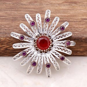 May include: A circular silver-toned brooch featuring a large, faceted red central stone surrounded by radiating filigree petals. Several smaller, round, deep red stones are set at intervals along the outer edges of the silver metal rays.
