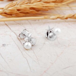 A pair of mini 925 sterling silver stud earrings featuring a detailed seashell and starfish motif accented with a small white pearl and butterfly backs.