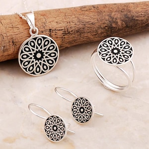 May include: A silver jewelry set consisting of a circular pendant on a chain, a matching ring with a split band, and two drop earrings. Each piece features a black circular inlay with a white geometric floral pattern radiating from the center.