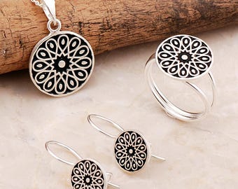 May include: A silver jewelry set consisting of a circular pendant on a chain, a matching ring with a split band, and two drop earrings. Each piece features a black circular inlay with a white geometric floral pattern radiating from the center.