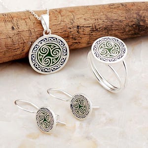 May include: A silver jewelry set consisting of a pendant on a chain, a ring, and a pair of drop earrings. Each piece features a circular design with a central green triskelion motif surrounded by a border of small, dark blue infinity symbols.