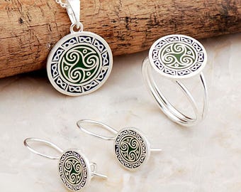 May include: A silver jewelry set consisting of a pendant on a chain, a ring, and a pair of drop earrings. Each piece features a circular design with a central green triskelion motif surrounded by a border of small, dark blue infinity symbols.