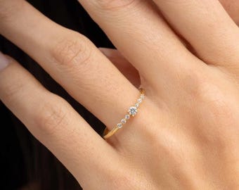May include: A thin gold band ring featuring a central round brilliant-cut diamond flanked by three smaller round diamonds on each side, arranged in a linear row along the top of the band, creating a delicate and refined jewelry piece.