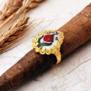 May include: A gold-toned ring featuring an oval-shaped center piece with a white background and a hand-painted red tulip design with green leaves. The metal setting has a scalloped, textured edge that frames the central floral motif.