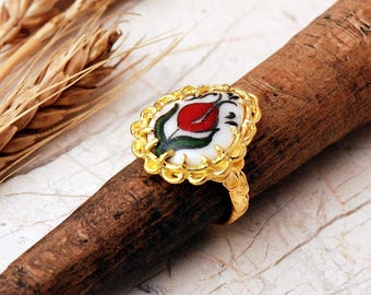 May include: A gold-toned ring featuring an oval-shaped center piece with a white background and a hand-painted red tulip design with green leaves. The metal setting has a scalloped, textured edge that frames the central floral motif.