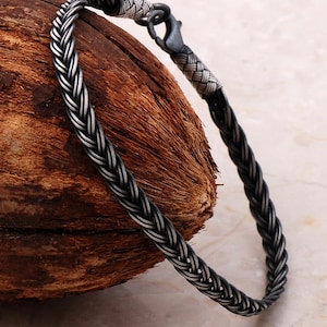 May include: A dark silver bracelet featuring a woven, braided chain design with a metallic finish. The bracelet includes a secure lobster claw clasp mechanism at the end, connecting the two ends of the intricate, rope-like metal link structure.