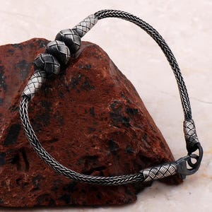 May include: A silver bracelet featuring a woven chain design with three spherical, textured beads positioned along the top section. The bracelet includes a secure metal clasp mechanism at one end, showcasing intricate craftsmanship and a polished finish.