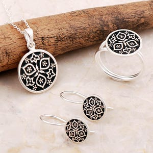 May include: A silver jewelry set consisting of a circular pendant on a chain, a ring, and a pair of drop earrings. Each piece features a black circular inlay with a white floral pattern consisting of four petals and small dots in the center.