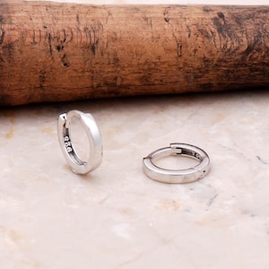 May include: A pair of small silver hoop earrings with a polished finish. Each earring features a hinged closure mechanism and is clearly stamped with the hallmark 925 on the inner surface, indicating sterling silver composition.