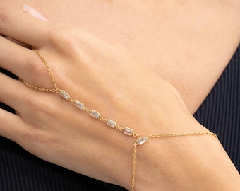 May include: A gold-toned hand chain bracelet featuring a delicate link chain that connects a ring loop to a wrist strap, accented with a series of small, clear, rectangular-cut stones spaced evenly along the central chain segment.