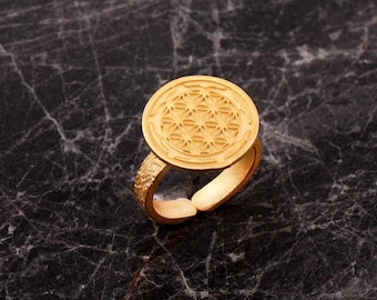 May include: A gold-toned ring featuring a circular face with an embossed geometric flower of life pattern. The band has an open, adjustable design with a textured surface on the sides, showcasing a matte finish throughout the piece.