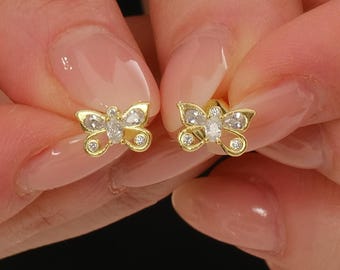 May include: A pair of gold butterfly-shaped stud earrings featuring clear, sparkling stones set within the wings. Each earring displays a central oval stone flanked by smaller round stones, creating a detailed insect silhouette in a polished gold finish.
