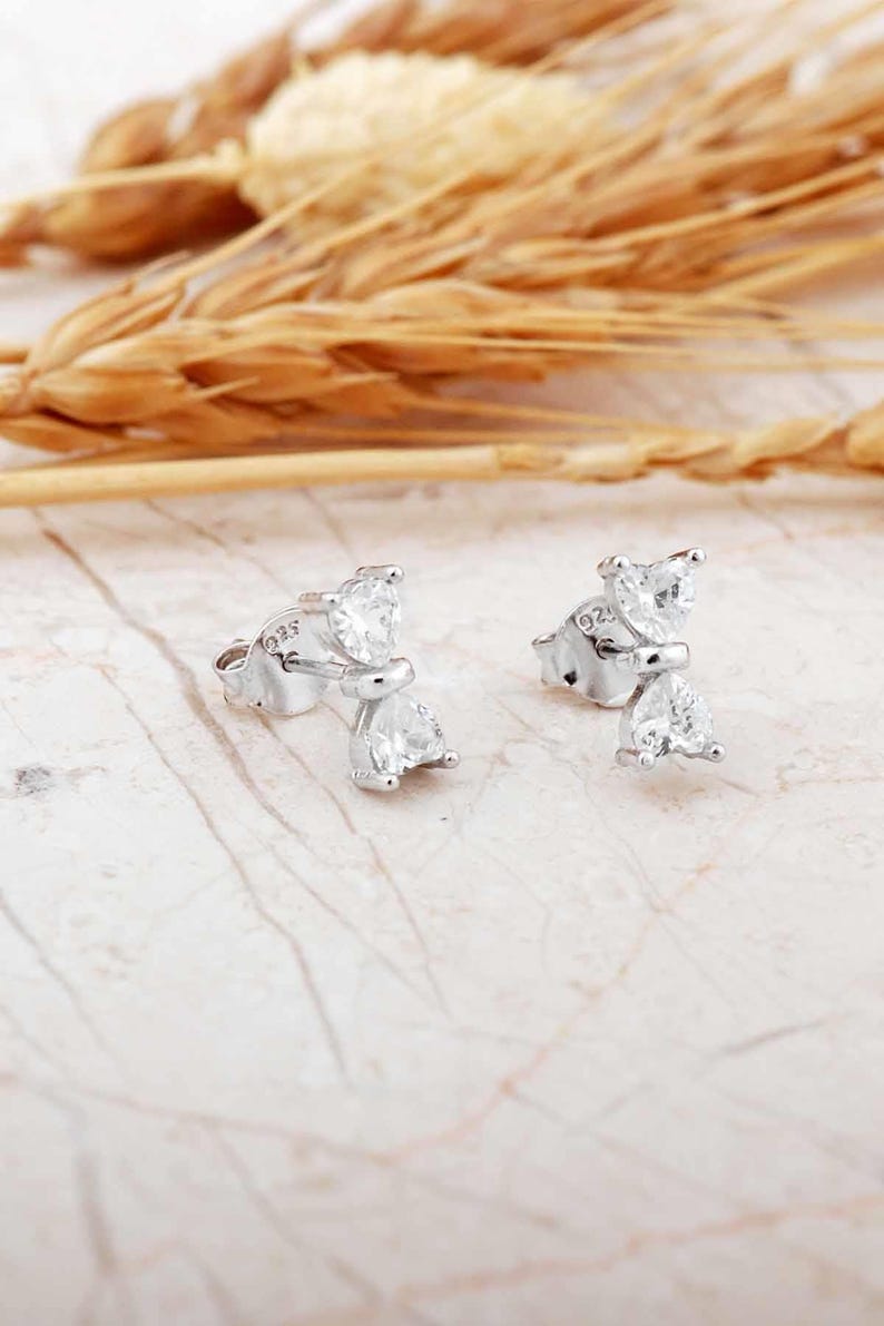 Mini bow‑shaped earrings made of rhodium‑plated sterling silver with a single white zircon stone on each side, measuring 4 mm tall by 1 cm wide.
