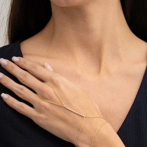 May include: A gold hand chain bracelet featuring a delicate chain that loops around one finger and connects to a horizontal bar set with small clear stones, extending to a thin chain that wraps around the wrist with a polished metallic finish.