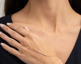 May include: A gold hand chain bracelet featuring a delicate chain that loops around one finger and connects to a horizontal bar set with small clear stones, extending to a thin chain that wraps around the wrist with a polished metallic finish.