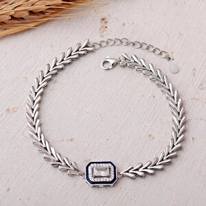 A detailed close-up of a 925 sterling silver herringbone chain bracelet featuring a dark blue enamel center piece adorned with a shimmering baguette-cut zircon stone.