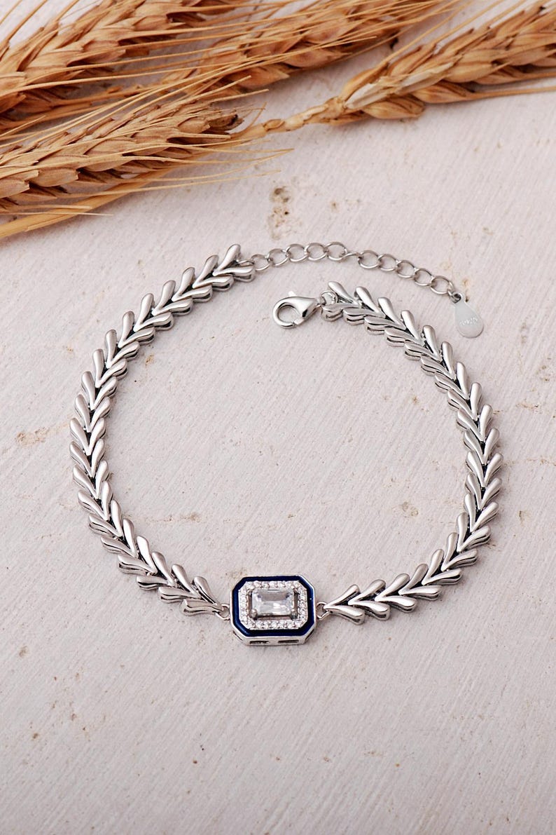A detailed close-up of a 925 sterling silver herringbone chain bracelet featuring a dark blue enamel center piece adorned with a shimmering baguette-cut zircon stone.