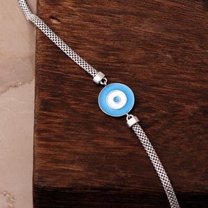 A silver bracelet with a blue Nazar evil‑eye bead in the center, white‑gold plated accents, and a parrot‑style clasp, displayed on a light background.