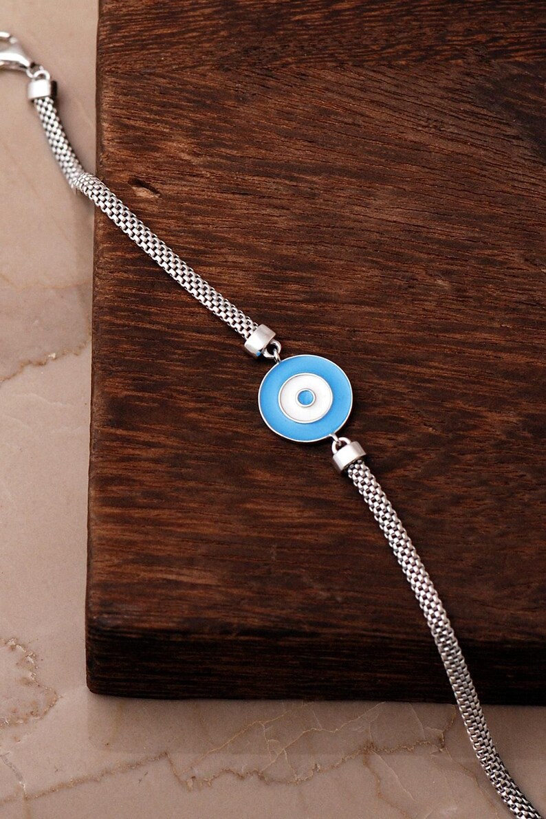 A silver bracelet with a blue Nazar evil‑eye bead in the center, white‑gold plated accents, and a parrot‑style clasp, displayed on a light background.
