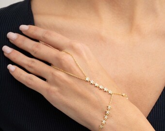 May include: A gold-toned hand chain featuring a delicate link design adorned with small, star-shaped charms. Each star is embedded with tiny, clear, sparkling stones. The chain connects a ring loop to a wrist bracelet, creating a continuous piece.