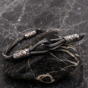 May include: A silver bracelet featuring a woven chain design with a central knot detail. The metal has an oxidized finish, creating a dark, textured appearance. The bracelet includes two decorative woven silver bands flanking the central knot.