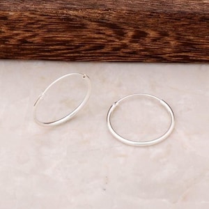May include: A pair of small, polished silver hoop earrings. Each earring features a thin, circular wire design with a simple hinged closure mechanism, showcasing a bright, reflective metallic finish that catches the light evenly across the surface.