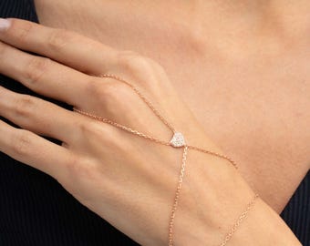 May include: A rose gold hand chain featuring a delicate metal link chain that connects a ring to a bracelet. The center of the hand chain is adorned with a small, heart-shaped charm encrusted with tiny, sparkling white stones.