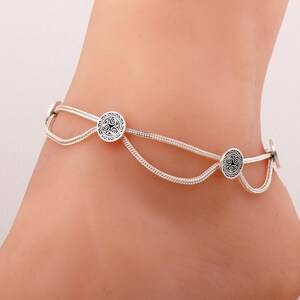 May include: A silver-coloured anklet featuring a double-strand chain design with two circular charms. Each charm displays a black triskelion symbol set against a silver background. The chain links are textured and connected by small metal loops.