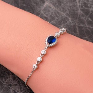 May include: A silver-toned chain bracelet featuring a central pear-shaped deep blue stone surrounded by a halo of small clear stones. The bracelet includes additional round clear stones set along the chain links on both sides of the central piece.