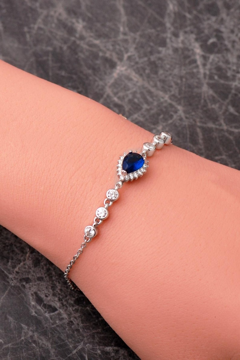 A rhodium‑plated 925 sterling silver bracelet with a dangling blue sapphire stone and sparkling cubic zirconia crystals, displayed on a soft white background.