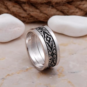 May include: A silver-toned metal ring featuring a wide band with a recessed center channel. The center channel contains a black, intricate floral and vine pattern that wraps around the entire circumference of the ring, contrasting with the smooth, polished silver edges.