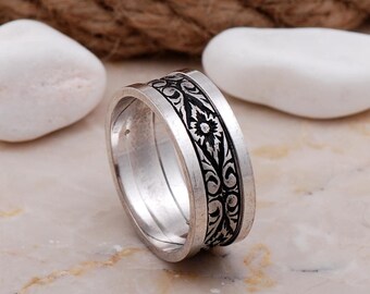 May include: A silver-toned metal ring featuring a wide band with a recessed center channel. The center channel contains a black, intricate floral and vine pattern that wraps around the entire circumference of the ring, contrasting with the smooth, polished silver edges.