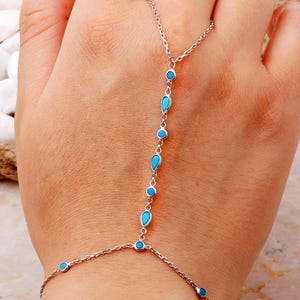 May include: A silver-toned hand chain bracelet featuring a delicate metal link chain adorned with small, alternating round and teardrop-shaped bright turquoise blue stones. The jewelry piece connects a wrist bracelet to a single finger ring.