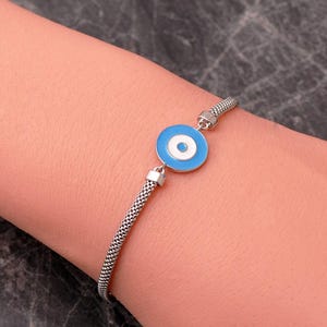 Op de afbeelding: Een zilverkleurige armband met een centrale ronde bedel met concentrische ringen in lichtblauw, wit en een kleiner lichtblauw midden. De armband is gemaakt van een fijne, getextureerde metalen mesh-ketting.