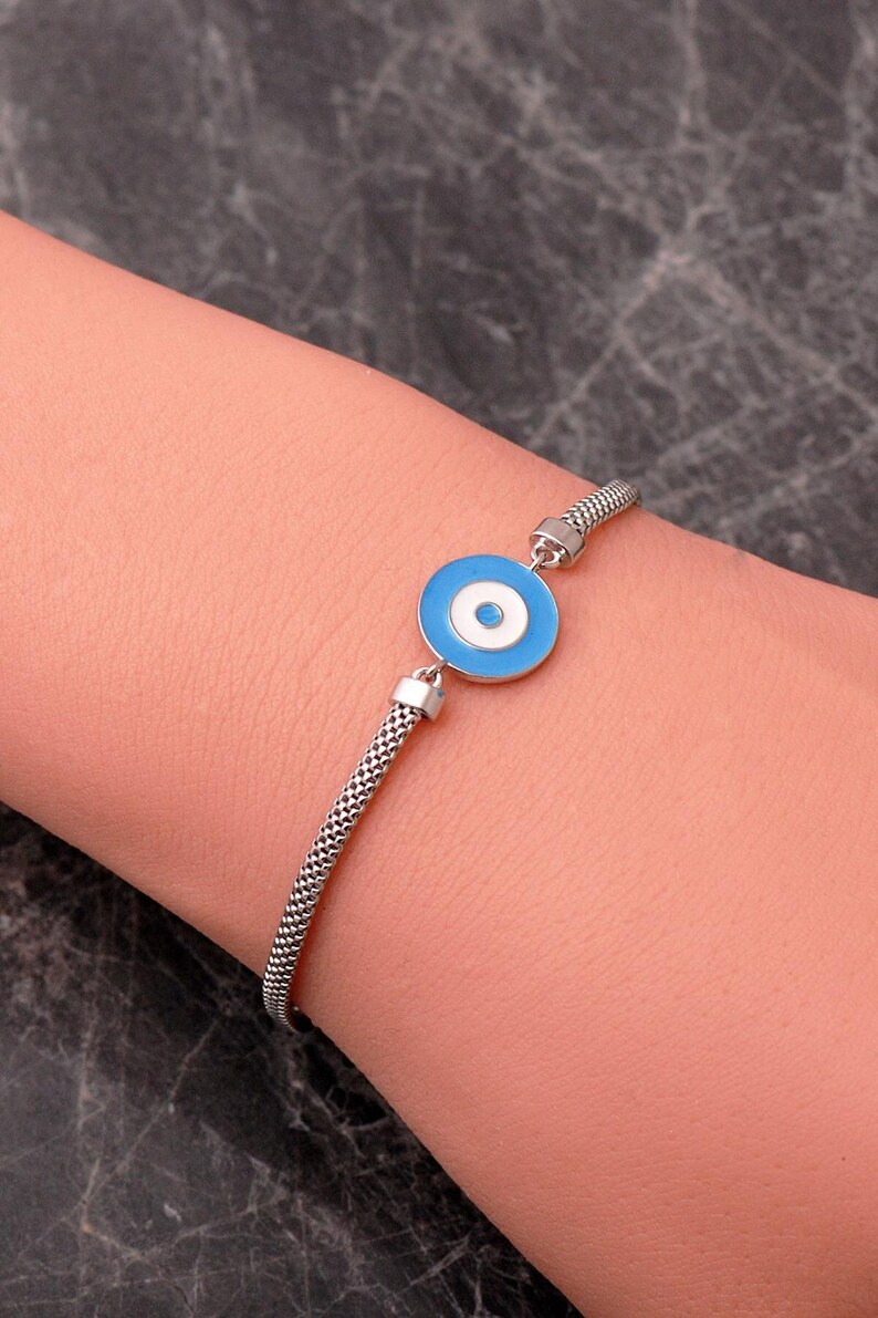 A silver bracelet with a blue Nazar evil‑eye bead in the center, white‑gold plated accents, and a parrot‑style clasp, displayed on a light background.