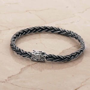 May include: A silver bracelet featuring a braided, rope-like chain design with a rectangular box clasp. The clasp is decorated with an engraved cross-hatch pattern. The metal has a dark, oxidized finish that highlights the intricate texture.