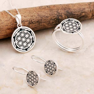 May include: A silver jewelry set consisting of a circular pendant on a chain, a ring, and a pair of drop earrings. Each piece features a black and silver circular inlay displaying a geometric flower of life pattern with intersecting circles.