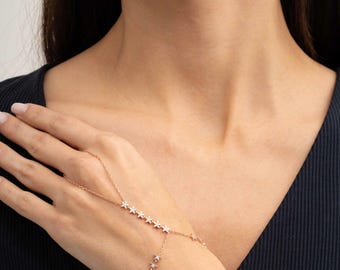 May include: A rose gold hand chain featuring a delicate link chain adorned with small, star-shaped charms. Each star is set with a tiny, clear, sparkling stone. The jewelry piece connects a ring component to a wrist chain, creating a continuous design.