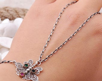 May include: A silver hand chain featuring a central butterfly motif with intricate filigree metalwork. The butterfly is adorned with three small, round, faceted gemstones in green, red, and black, connected by a delicate silver link chain.
