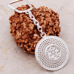 May include: Silver key chain with a round, intricate, silver charm. The charm has a woven design with a floral pattern in the center.