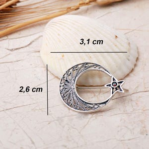 An intricate handmade 925 sterling silver brooch featuring filigree wirework in a crescent moon and star motif, set with a small round red garnet stone and finished with an oxidized antique silver coating.