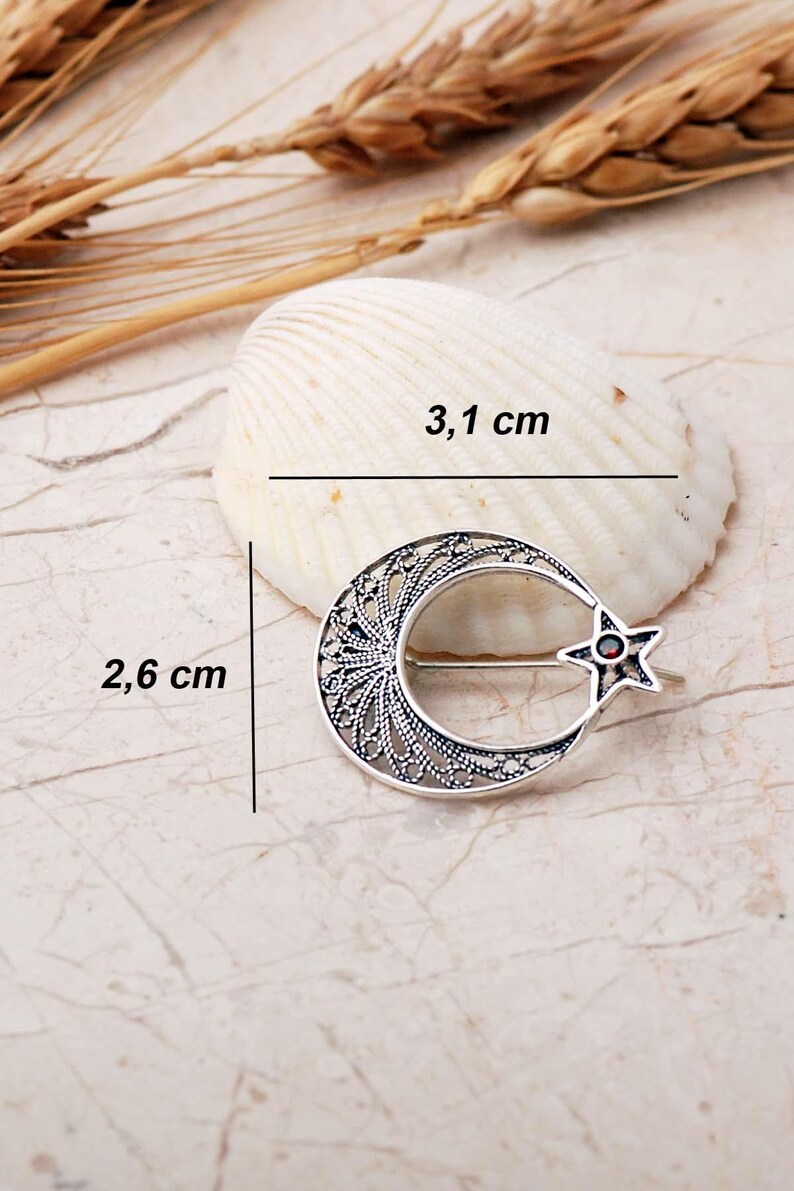 An intricate handmade 925 sterling silver brooch featuring filigree wirework in a crescent moon and star motif, set with a small round red garnet stone and finished with an oxidized antique silver coating.
