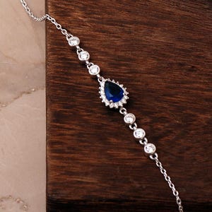 A rhodium‑plated 925 sterling silver bracelet with a dangling blue sapphire stone and sparkling cubic zirconia crystals, displayed on a soft white background.