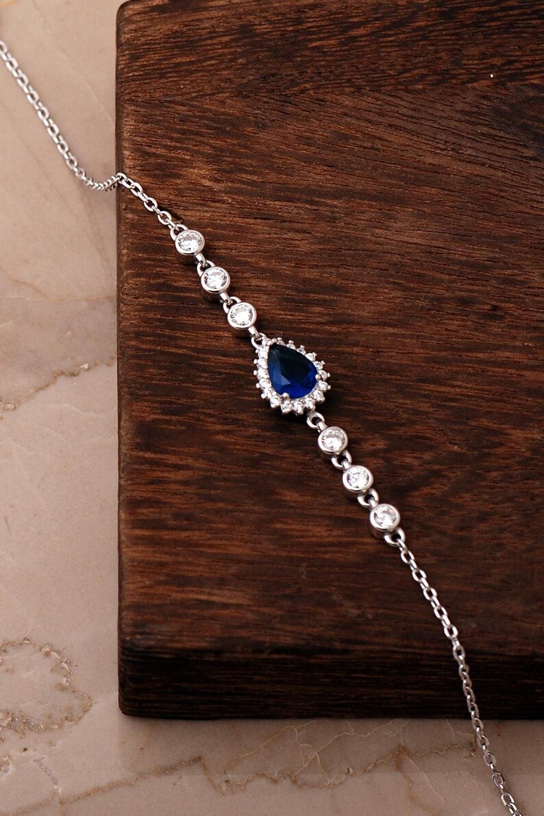 A rhodium‑plated 925 sterling silver bracelet with a dangling blue sapphire stone and sparkling cubic zirconia crystals, displayed on a soft white background.