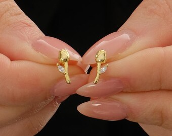 May include: A pair of gold-toned stud earrings shaped like small tulips. Each earring features a smooth, polished gold flower bud and a single, clear, marquise-cut gemstone accent set on the stem, creating a delicate and refined appearance.