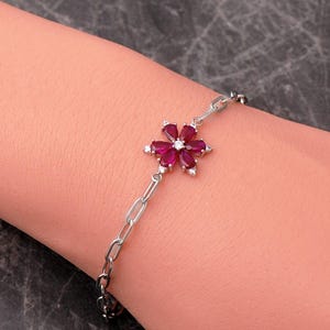 May include: A silver-toned chain bracelet featuring a central flower-shaped charm. The charm is composed of six pear-cut deep red stones arranged as petals around a small clear center stone, with additional clear stones accenting the flower base.