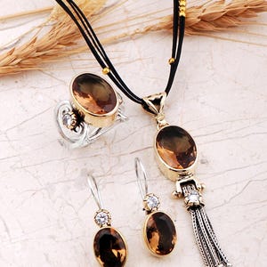 May include: A jewelry set consisting of a necklace, a ring, and a pair of earrings. Each piece features oval-cut brown gemstones set in gold-tone metal with silver-tone accents. The necklace includes a tassel pendant on a black cord.