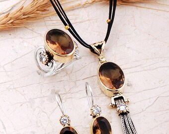 May include: A jewelry set consisting of a necklace, a ring, and a pair of earrings. Each piece features oval-cut brown gemstones set in gold-tone metal with silver-tone accents. The necklace includes a tassel pendant on a black cord.